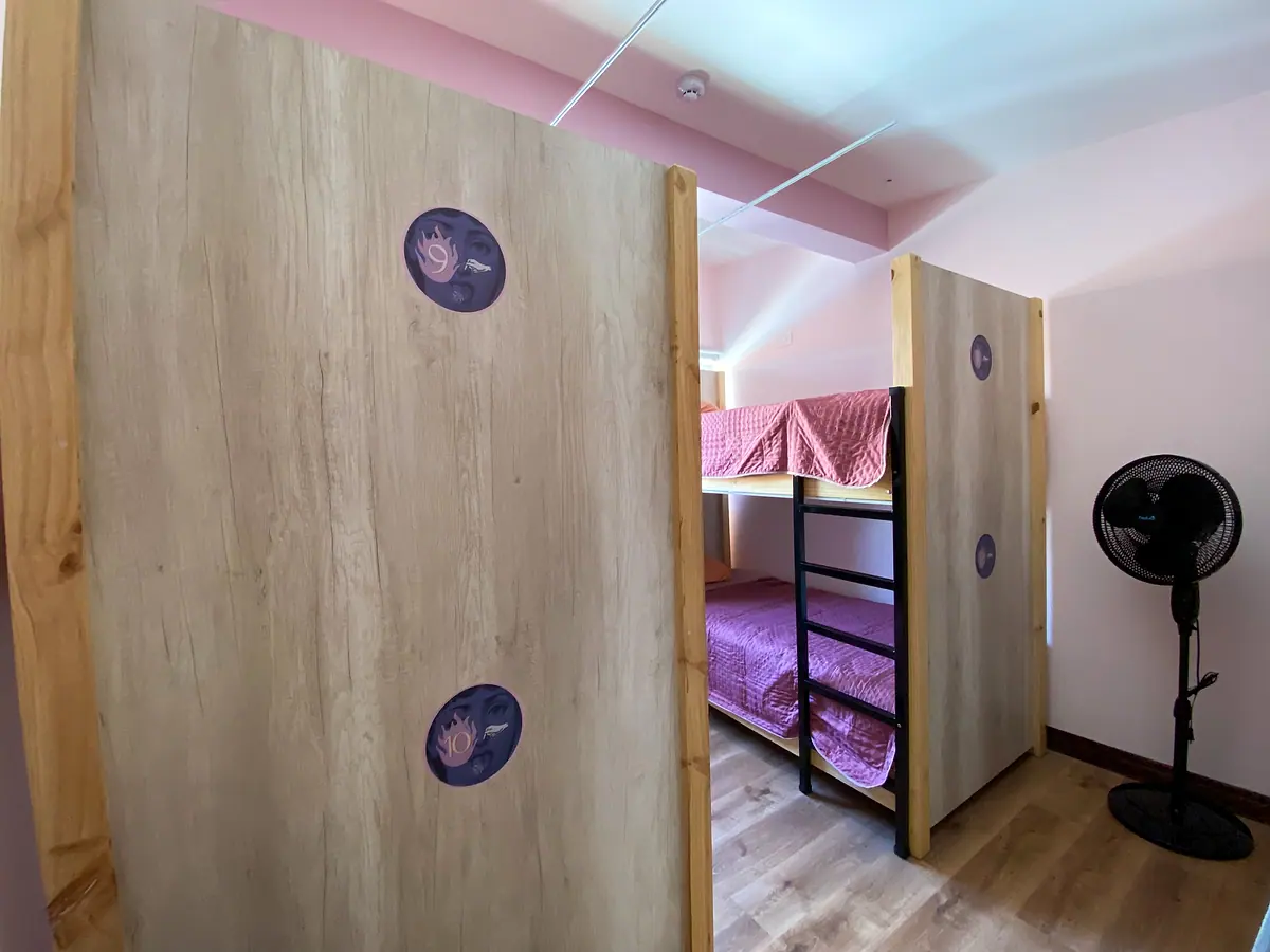 Fuego dorm: wooden partitions with Casa Pandora stickers, a bunk bed with pink bedding and a standing fan
