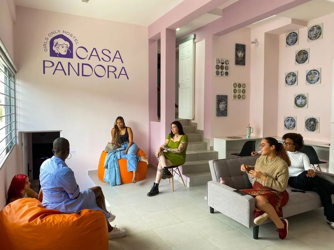 Women chatting in the pink lounge under the violet Casa Pandora wall logo, two of them on orange beanbags