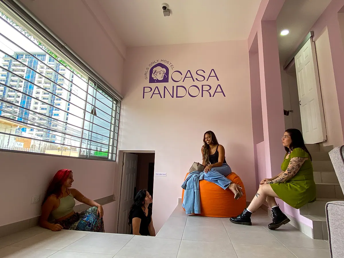 The pink lounge under the violet “Girls Only Hostel · Casa Pandora” wall logo: one guest on an orange beanbag, others on the steps and a chair