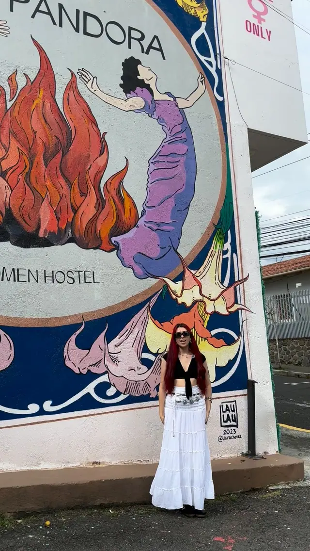 A woman in a white dress stands in front of the painted Casa Pandora mural: women in purple dancing around orange flames