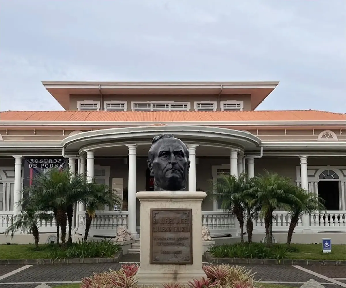 Museo Dr. Rafael Ángel Calderón Guardia: a white colonnaded house with an orange roof and a bronze bust in the garden